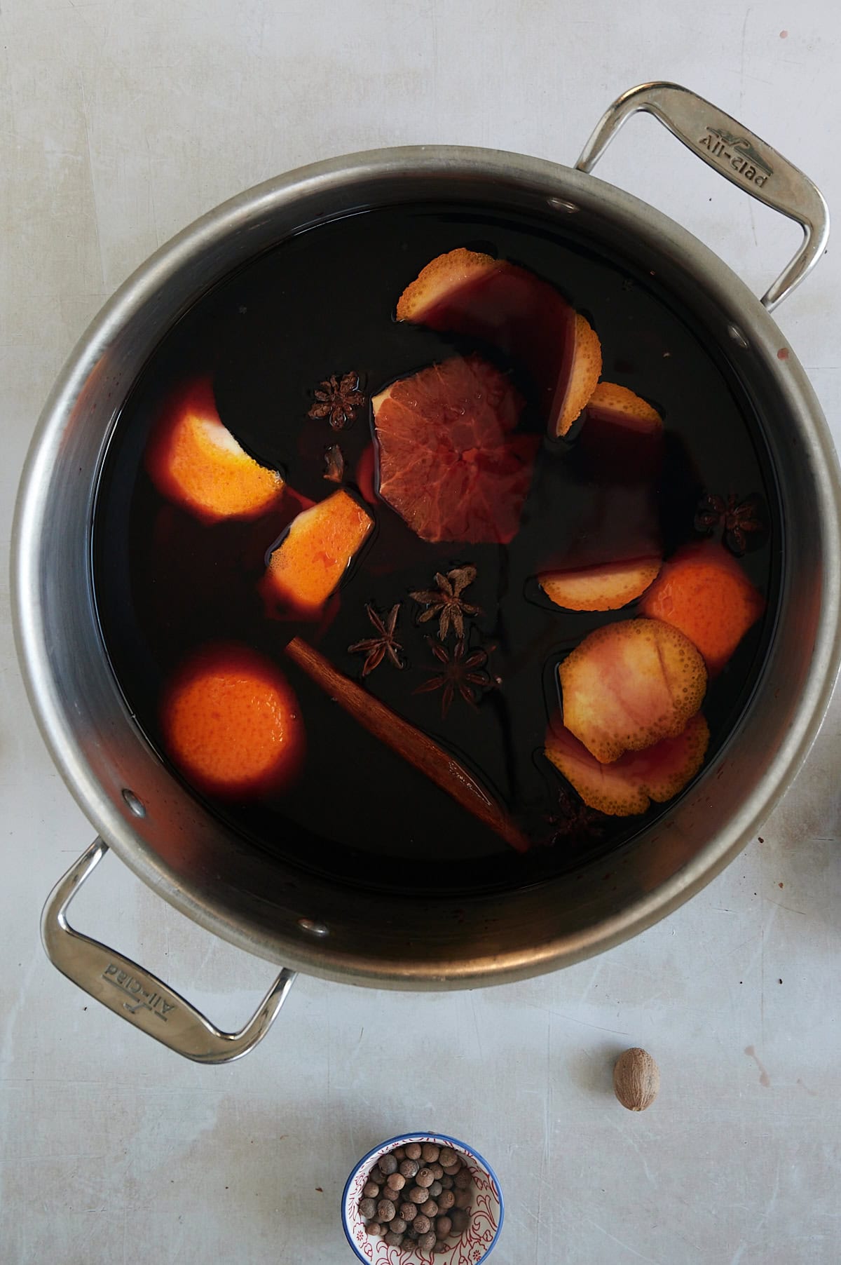 simmering pot of mulled red wine with orange peels, star anise, oranges and cinnamon sticks with bowls of whole cloves.