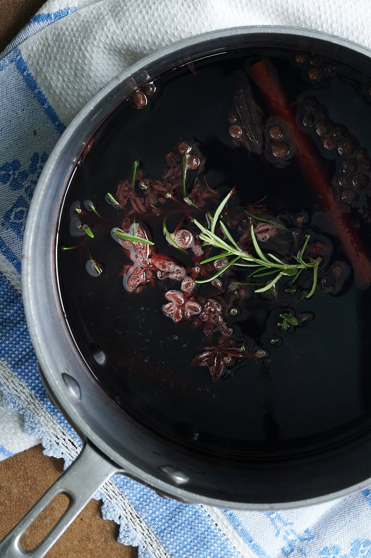 pot of mulled wine with rosemary, star anise, whole cloves and cinnamon sticks on a blue and white dish towel.