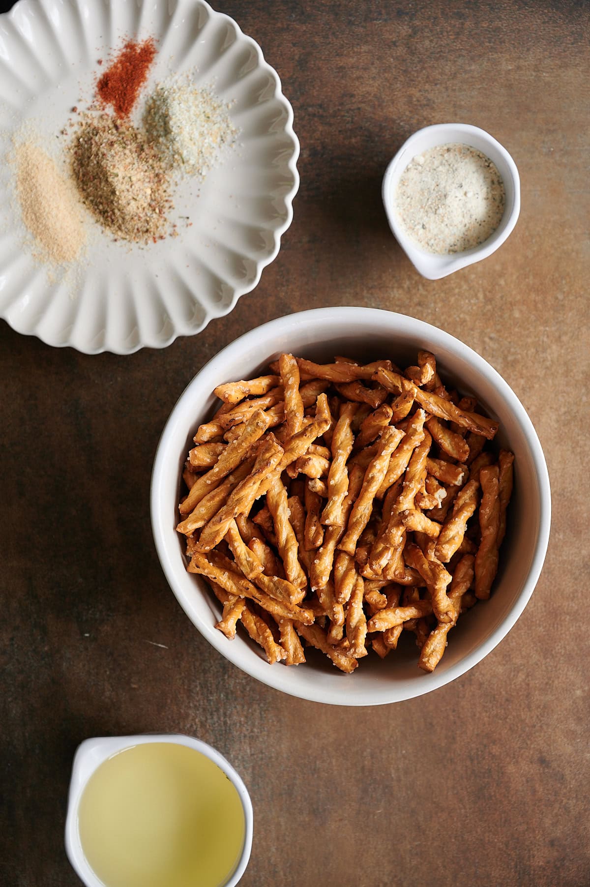 Ingredients for parmesan garlic pretzels in white bowls and plates including braided pretzels, seasoning and small bowl of oil.