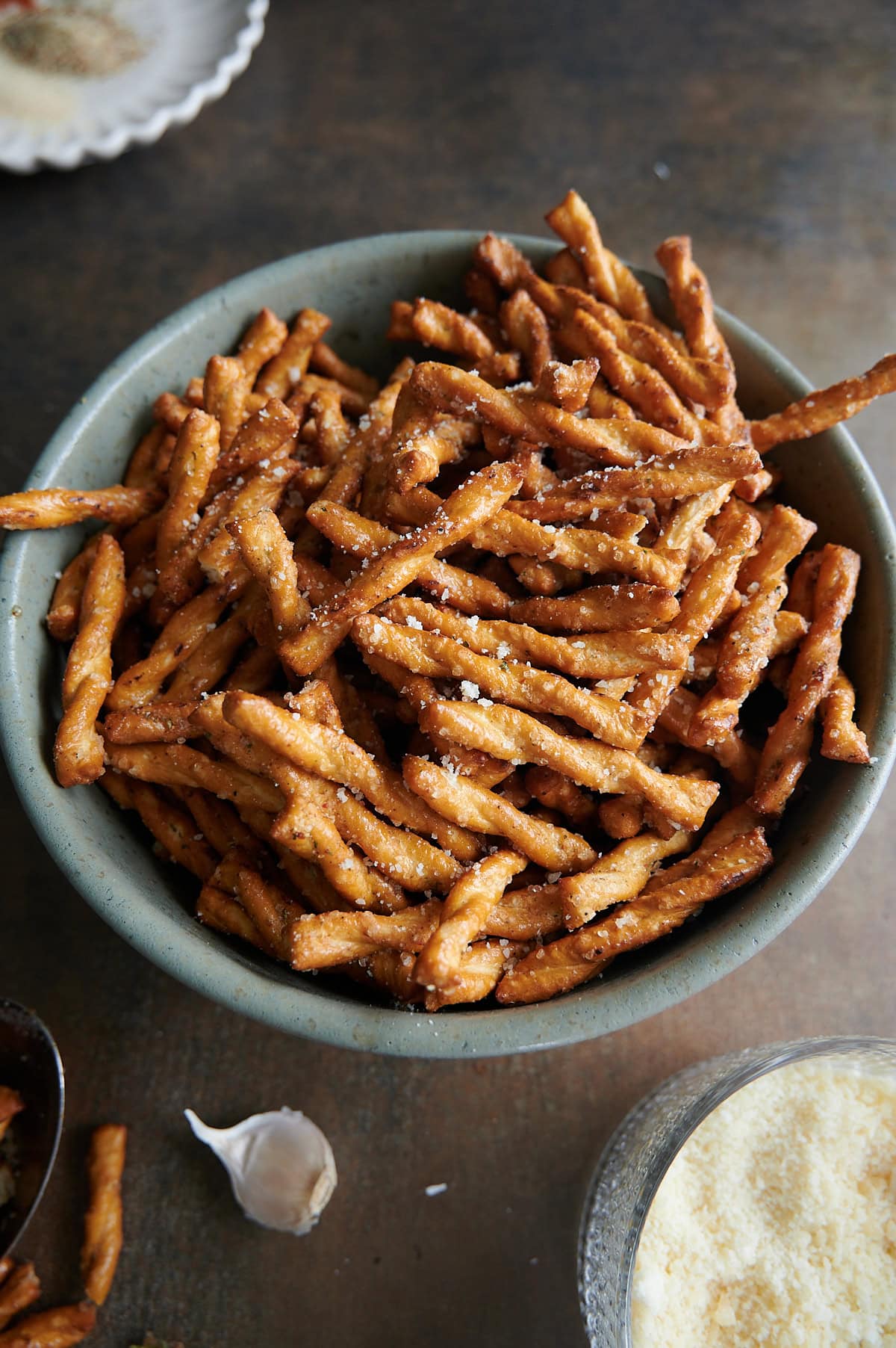 A green bowl of garlic parmesan pretzels with fresh garlic and bowl of parmesan on a dark brown background.
