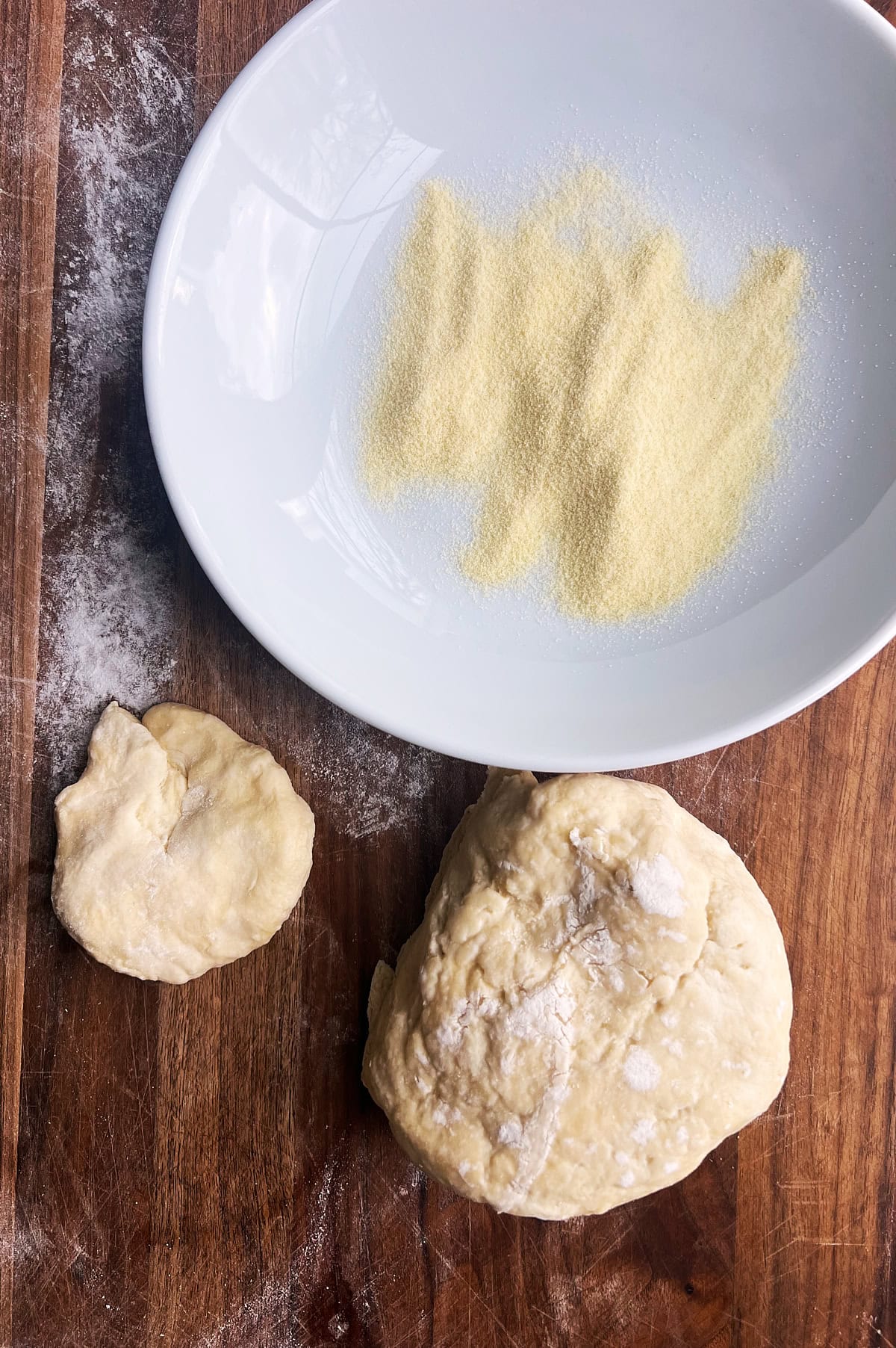 A piece of pasta dough cut of a ball of homemade pasta dough with a white bowl of semolina flour.