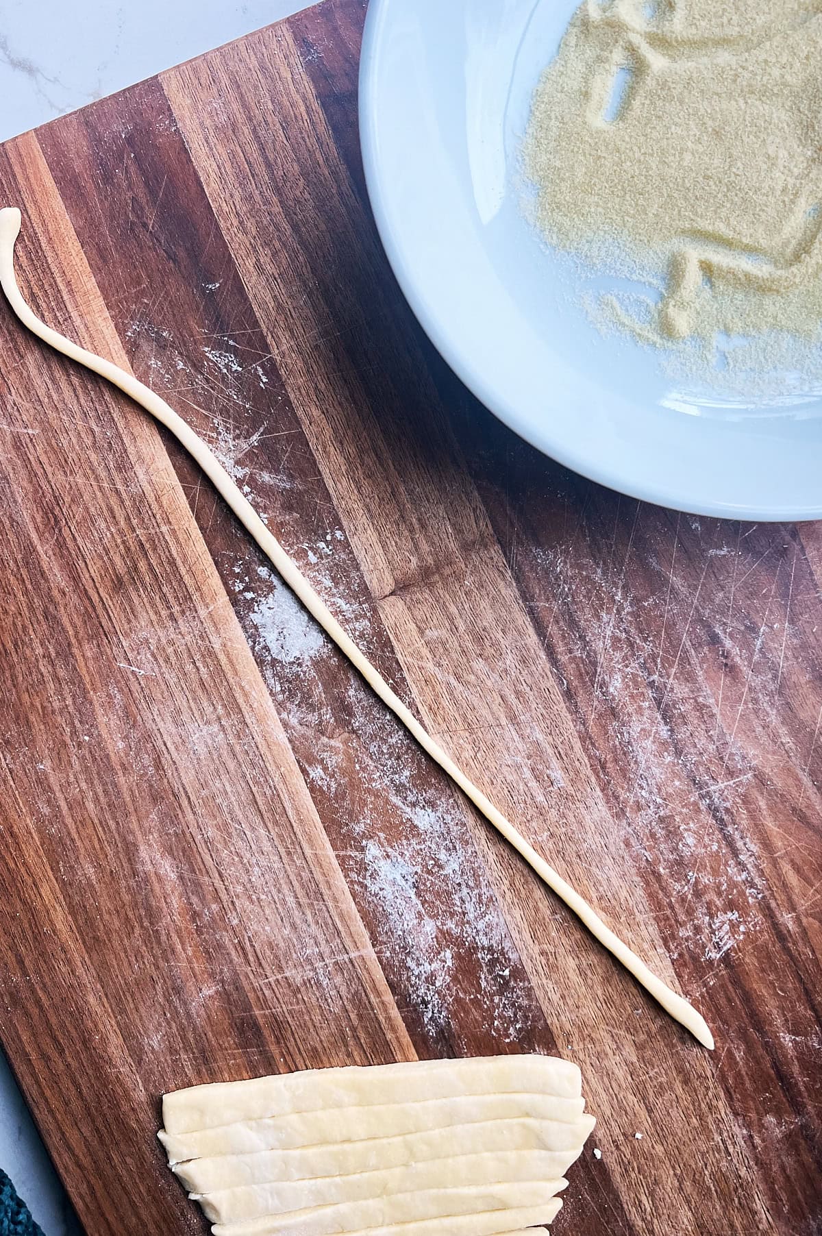 a rolled pici noodle on a wooden board with cut dough nearby.