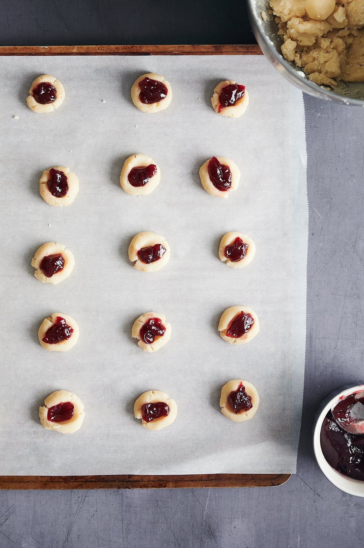 Raspberry jam filled thumbprint cookies on parchment paper covered baking sheet with bowl of jam.