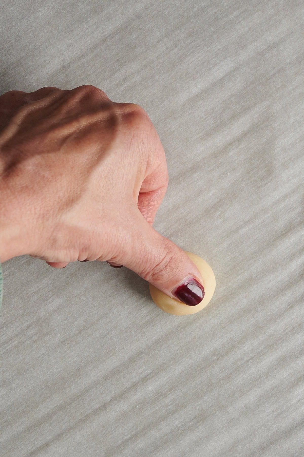 Hand using thumb to make indentation in cookie dough on parchment paper for thumbprint cookies.