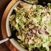 Overhead view of wood trimmed green large bowl of shaved Brussels sprouts with almonds, dates, parmesan and black serving utensils.