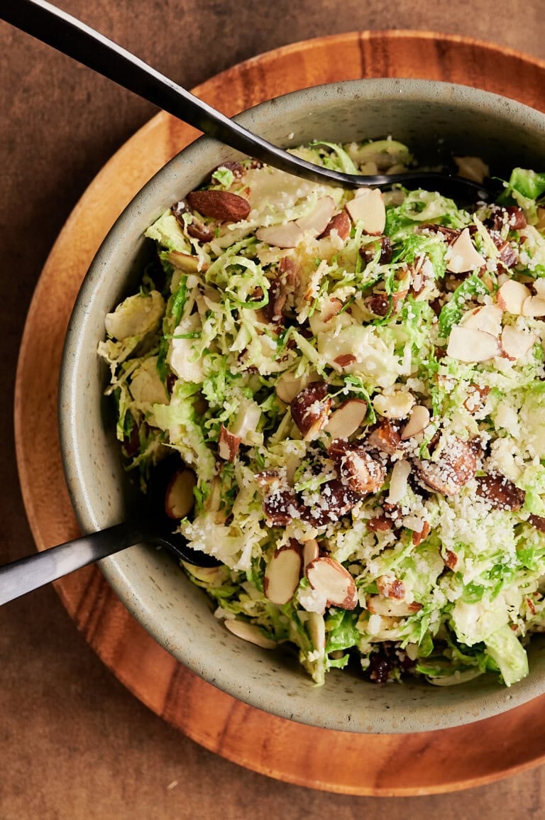 Overhead view of wood trimmed green large bowl of shaved Brussels sprouts with almonds, dates, parmesan and black serving utensils.