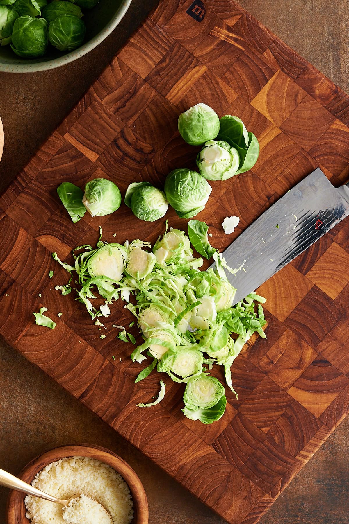Wooden cutting board with thinly sliced Brussels sprouts with a knife and bowls of Brussels sprouts and cheese nearby.
