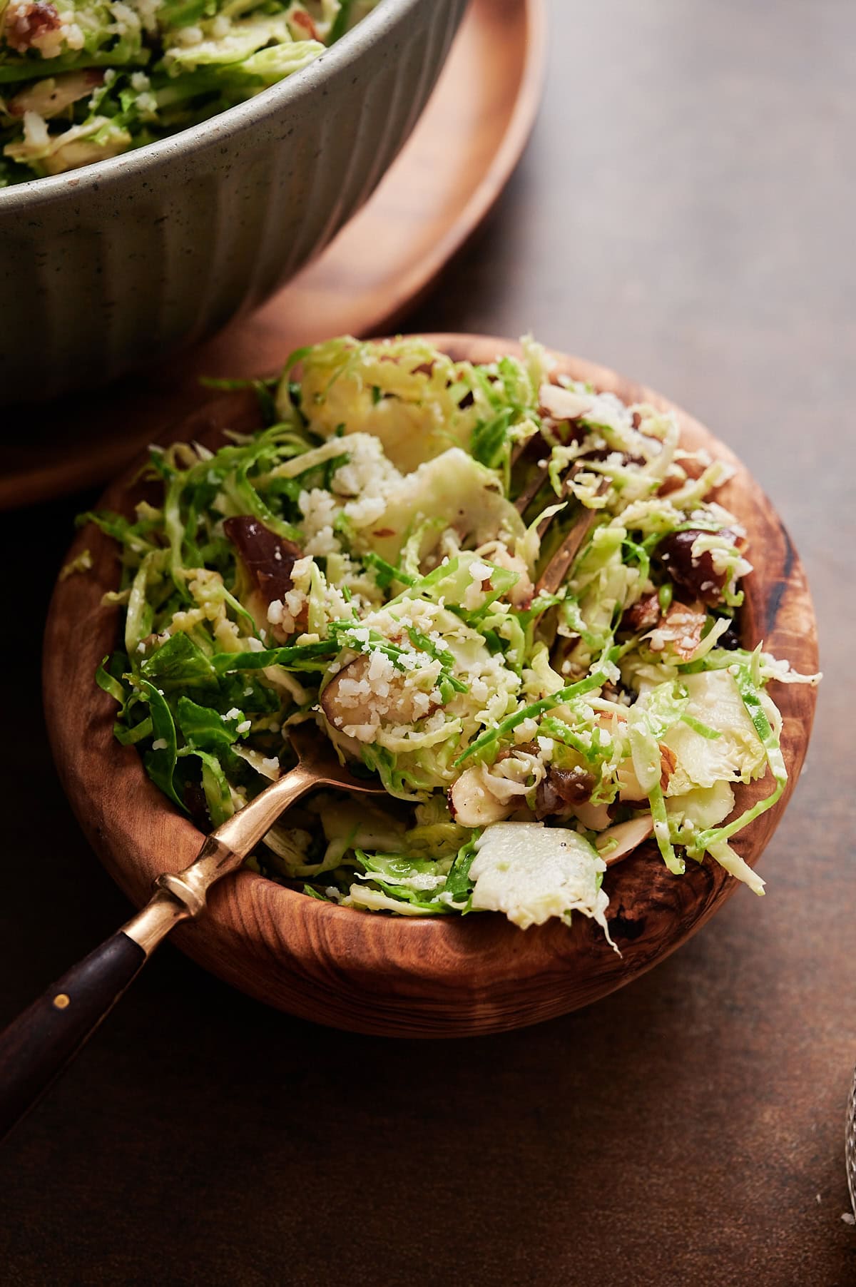 small wooden bowl with shredded Brussels sprouts, dates and parmesan cheese with an angled spoon and bowl of salad behind.