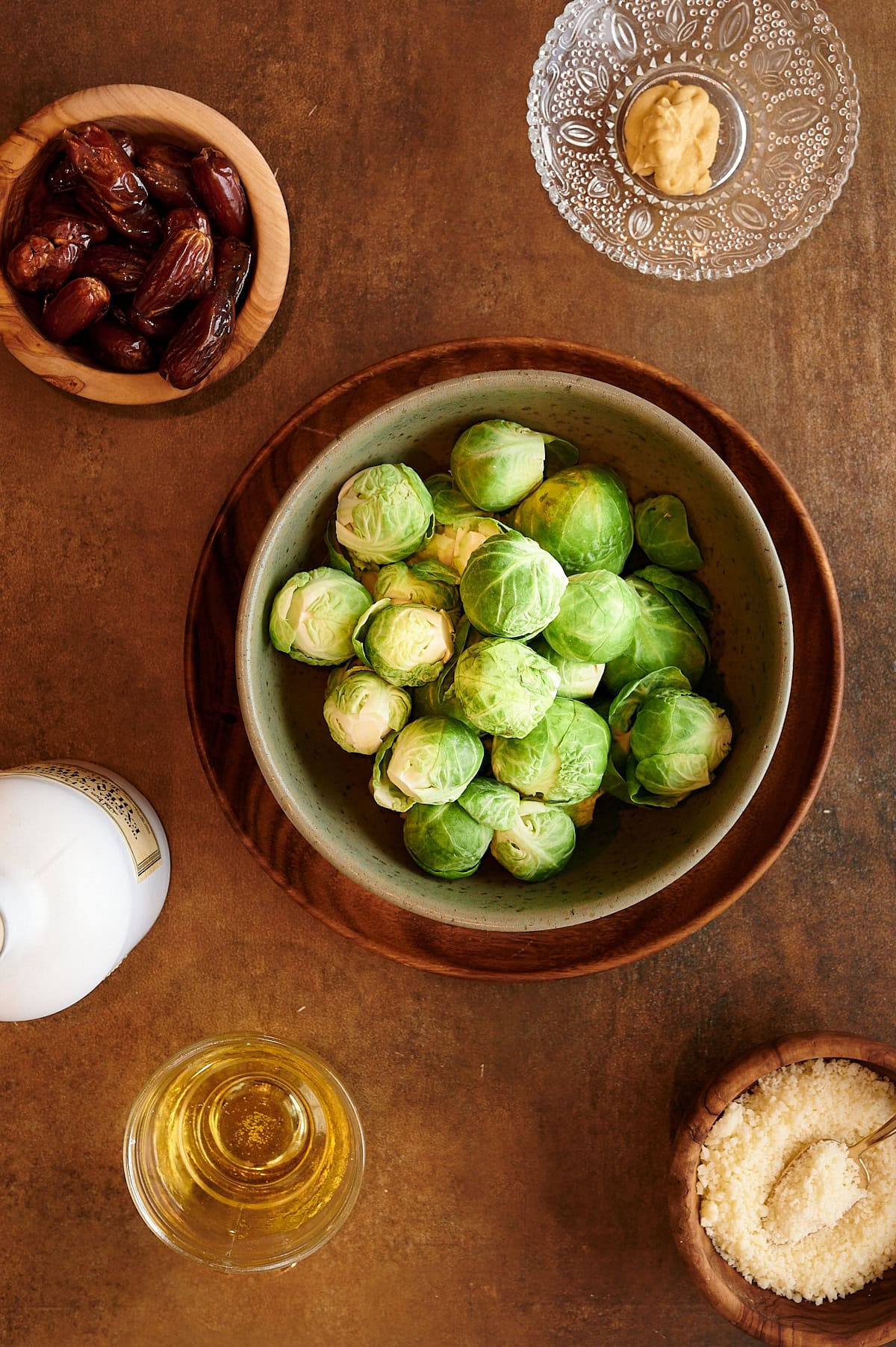 Ingredients for Brussels sprouts shredded salad in a green bowl with bowls of dates, Dijon mustard, parmesan cheese, vinegar and olive oil on a brown background.