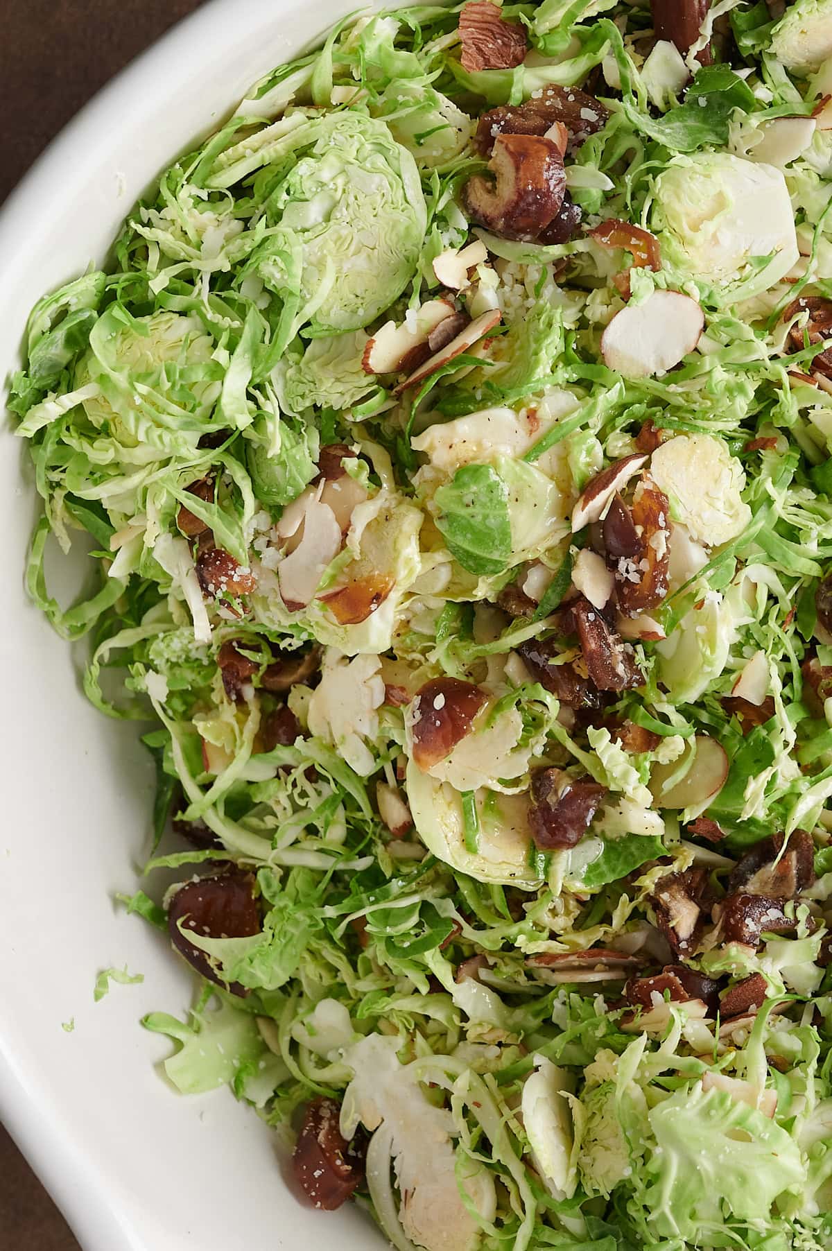 White bowl of Brussels sprouts shaved and tossed with almonds, cheese and dates.