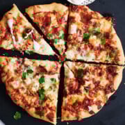 Whole chicken bacon pizza cut into six pieces on a black pizza peel with bowl of ranch dressing.