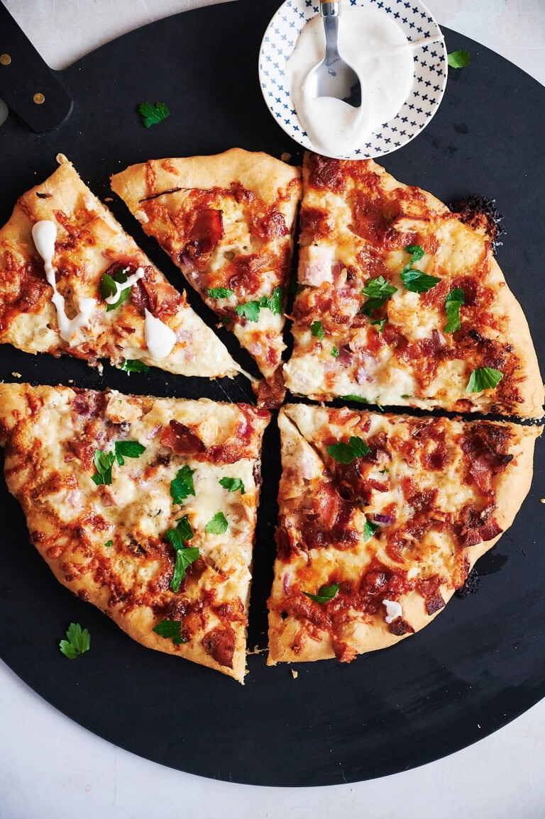 Whole chicken bacon pizza cut into six pieces on a black pizza peel with bowl of ranch dressing.
