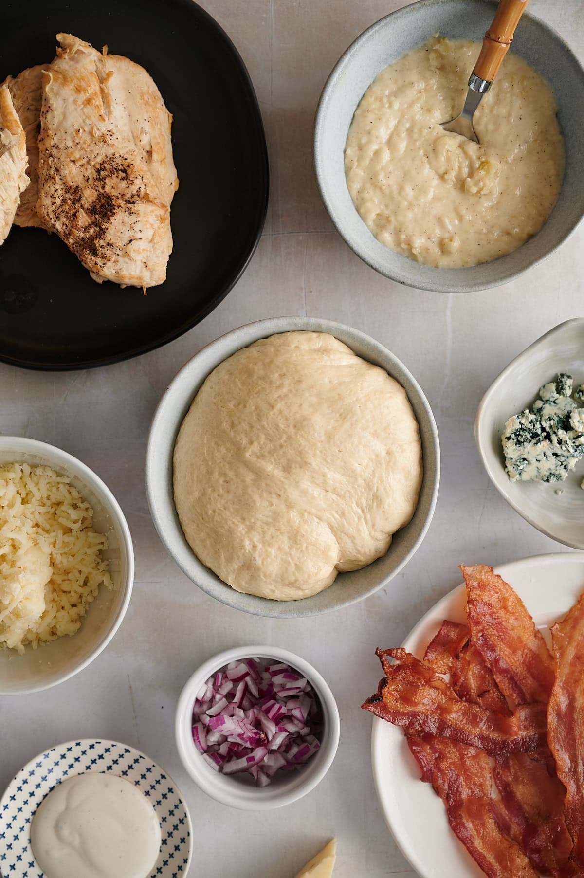 Ingredients for chicken bacon ranch pizza in bowls including pizza dough, sauce, chicken, bacon, cheeses and ranch dressing.