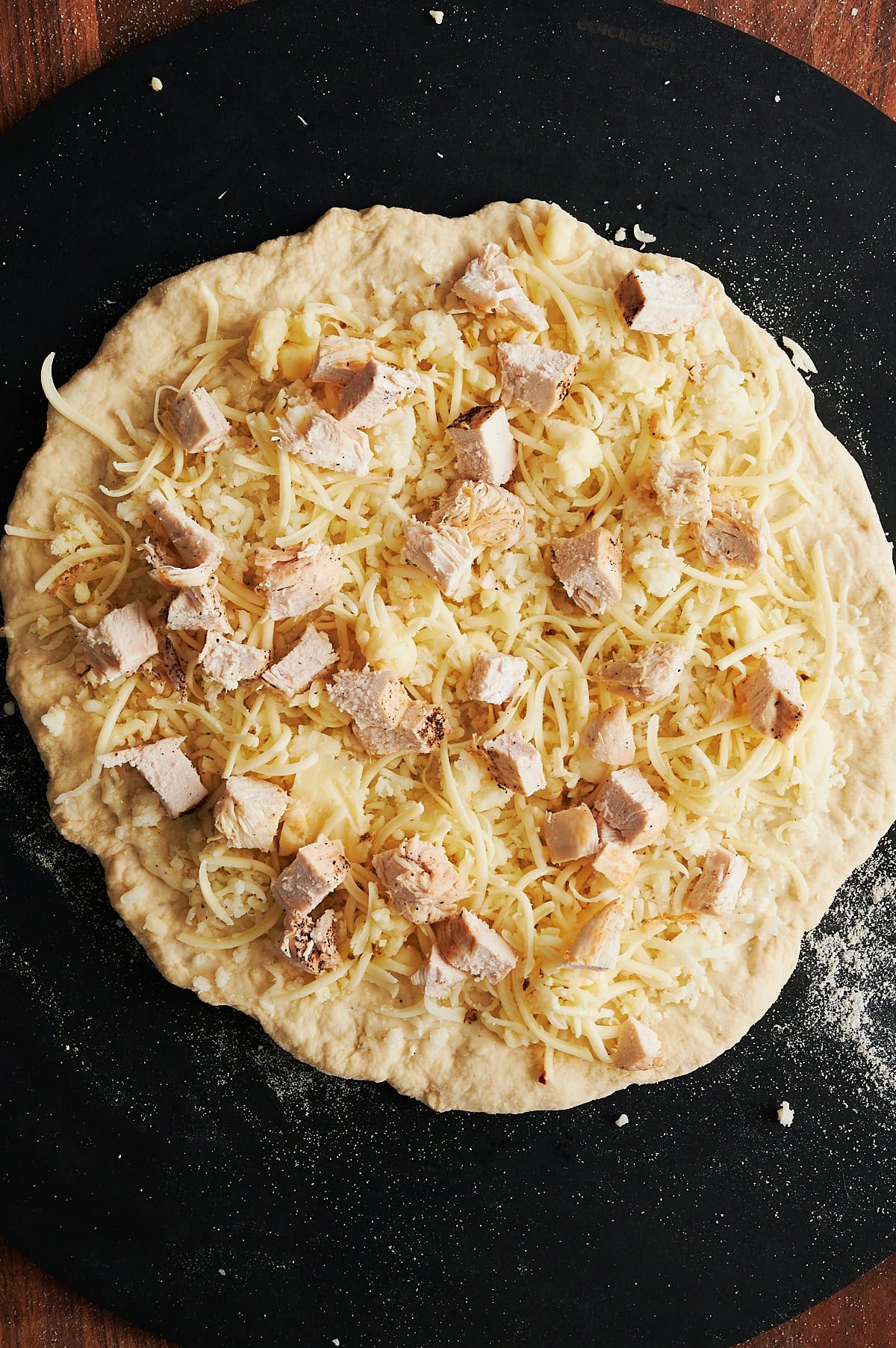 Pizza dough covered with cheese and chicken on a black pizza peel sprinkled with cornmeal.