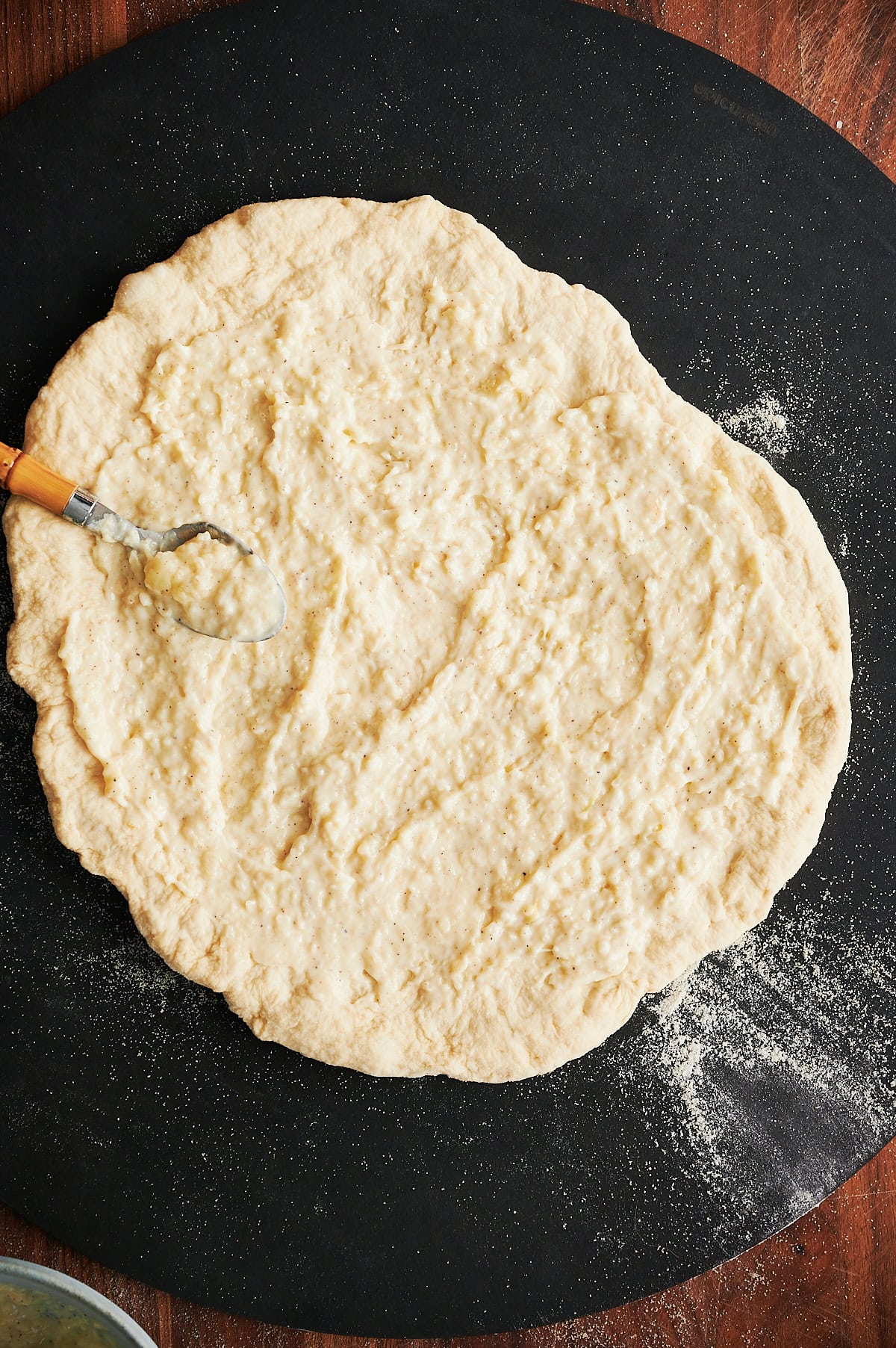 Creamy white pizza sauce being spread on pizza dough on a black pizza peel.