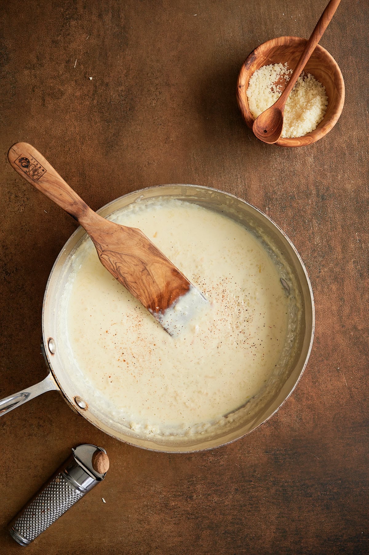 Grated nutmeg being added to stainless skillet of roasted garlic cream pizza sauce with wooden bowl of parmesan.