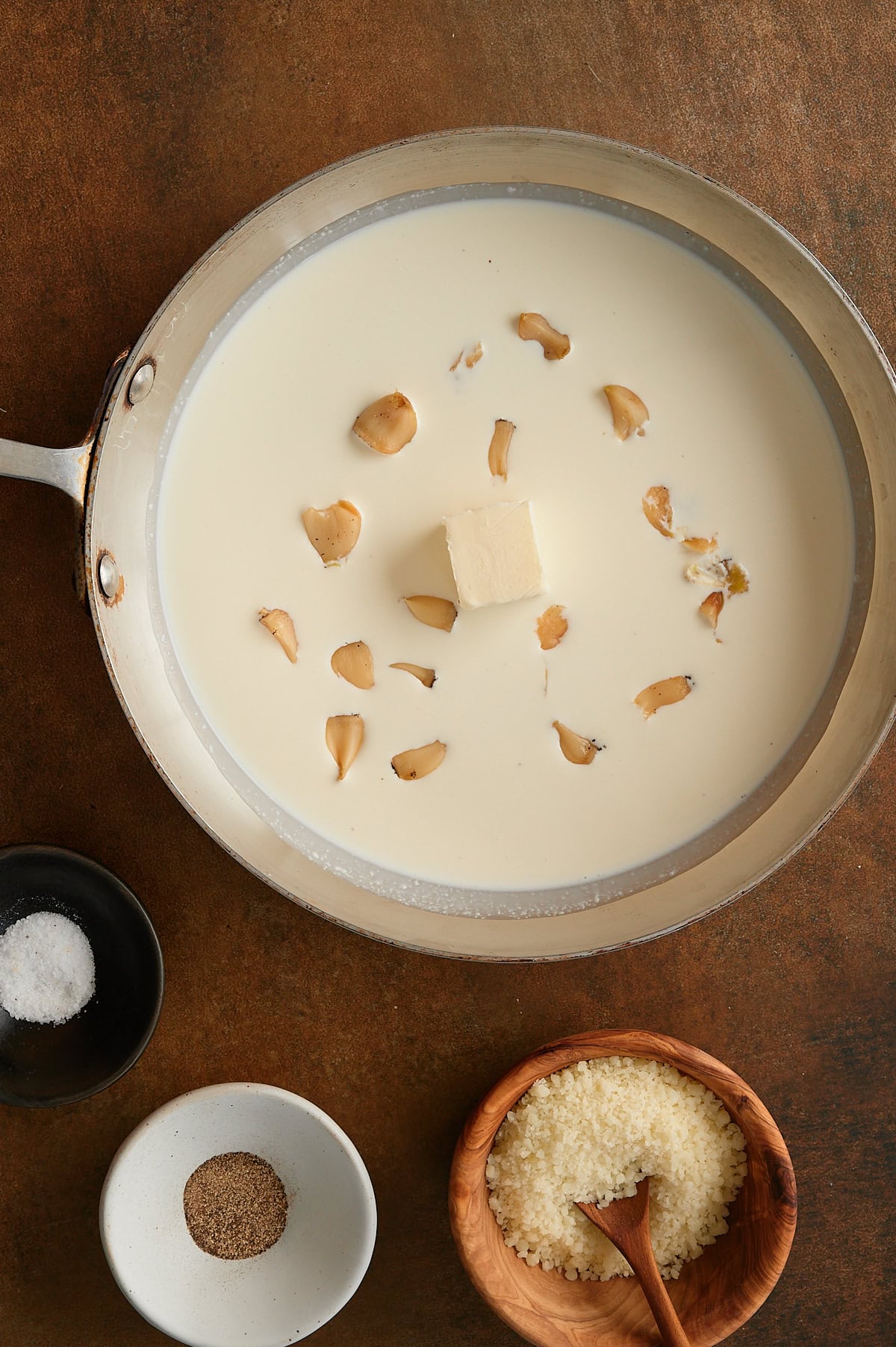 Cream in a skillet sprinkled with roasted garlic cloves and bowls of kosher salt, pepper and parmesan on a brown background.
