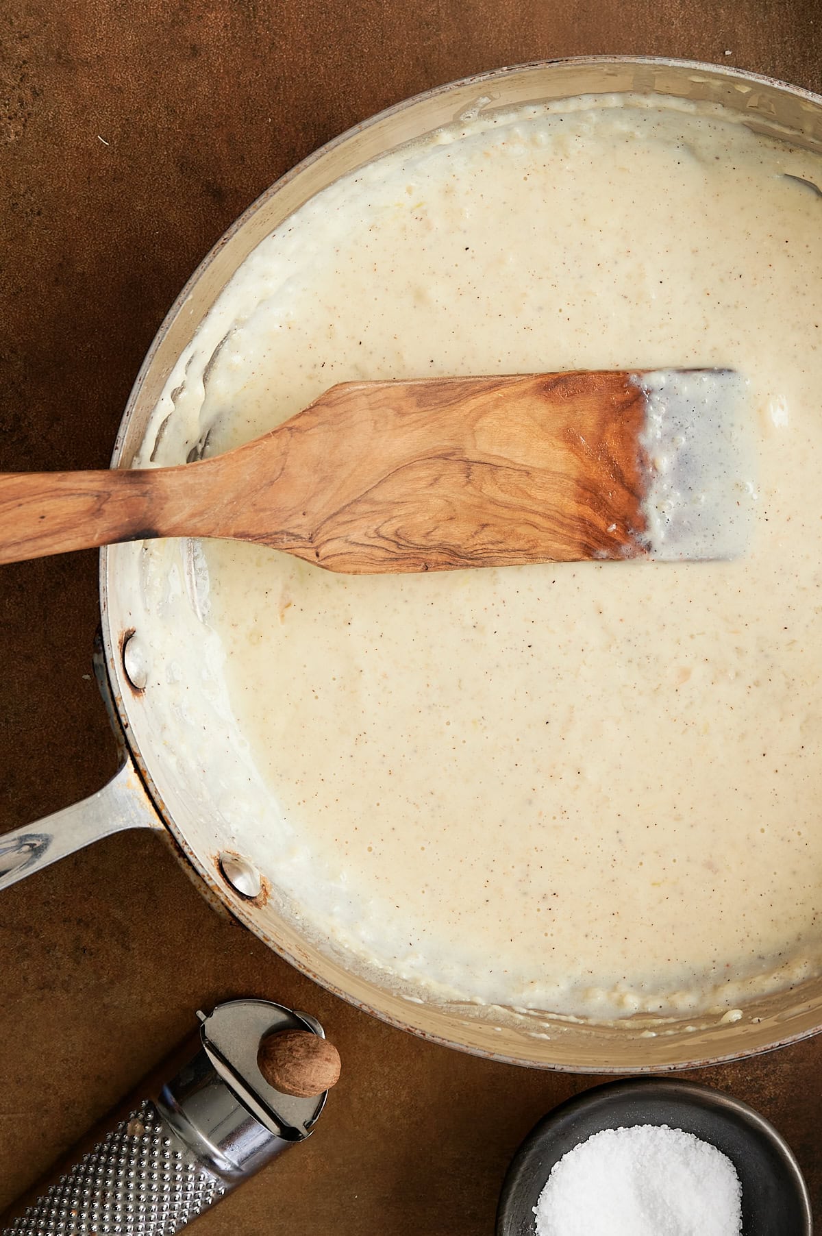 Skillet of roasted garlic pizza sauce with a wooden paddle, stainless grater of whole nutmeg and black bowl of kosher salt.