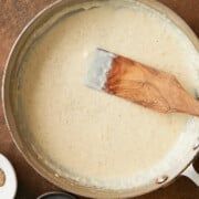 Finished stainless skillet of garlic pizza sauce with a wooden paddle and bowls of parmesan, salt, pepper and grated fresh nutmeg.