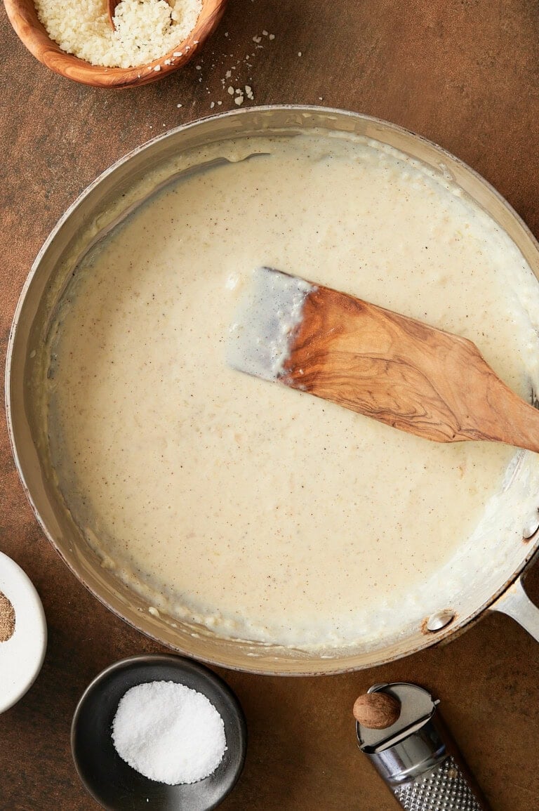 Finished stainless skillet of garlic pizza sauce with a wooden paddle and bowls of parmesan, salt, pepper and grated fresh nutmeg.