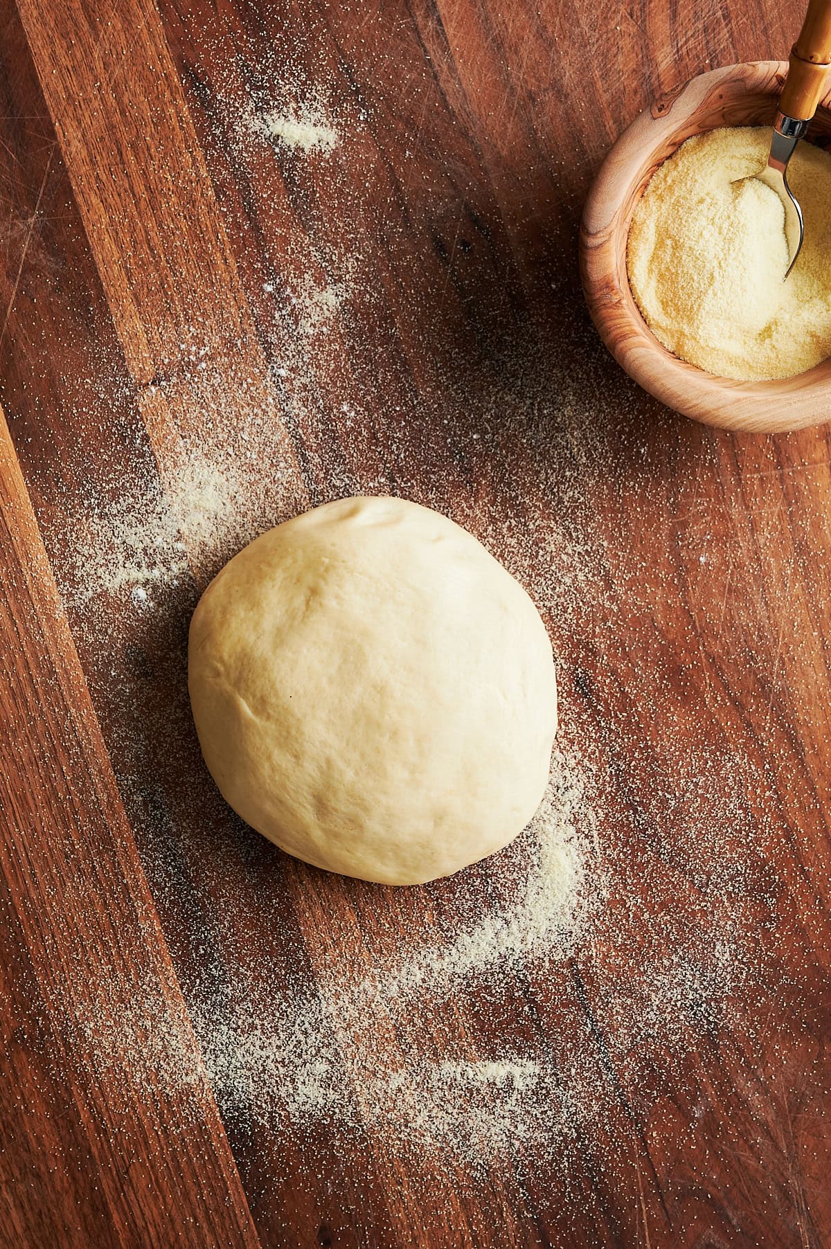 Ball of pizza dough on a wooden board sprinkled with semolina flour and wooden bowl of semolina flour nearby.