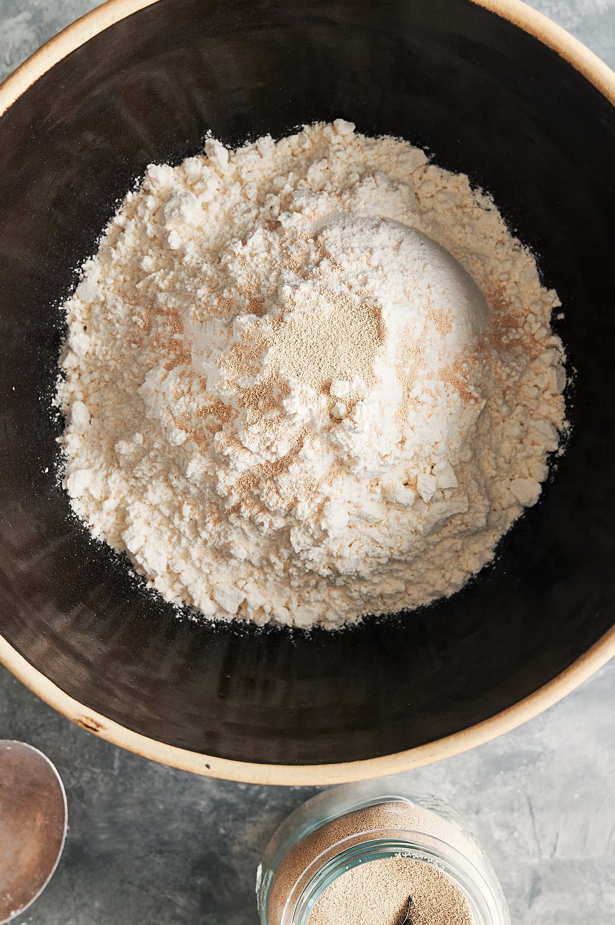 Black bowl of flour and yeast for pizza dough and jar of yeast nearby.