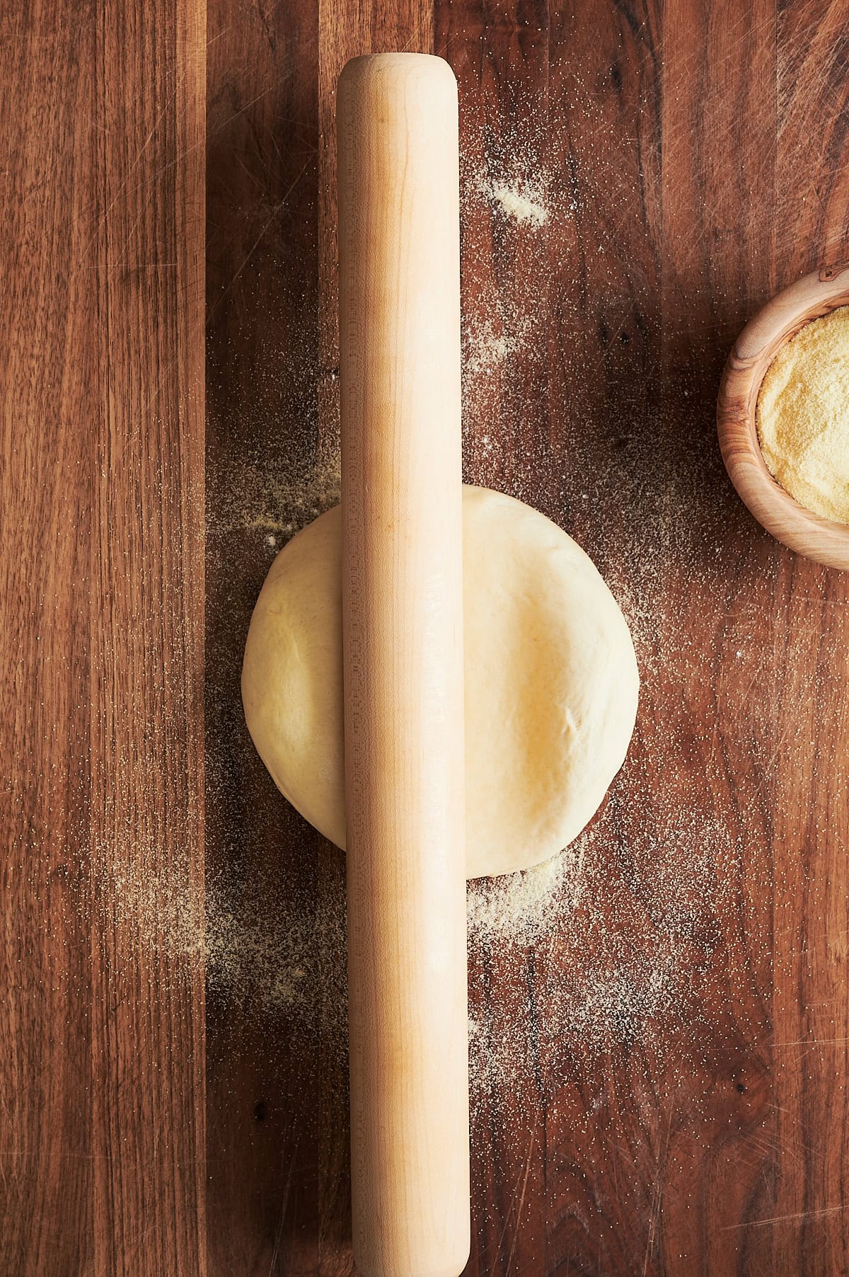 Thin crust pizza in a ball starting to being rolled out by a French rolling pin on a wooden cutting board.