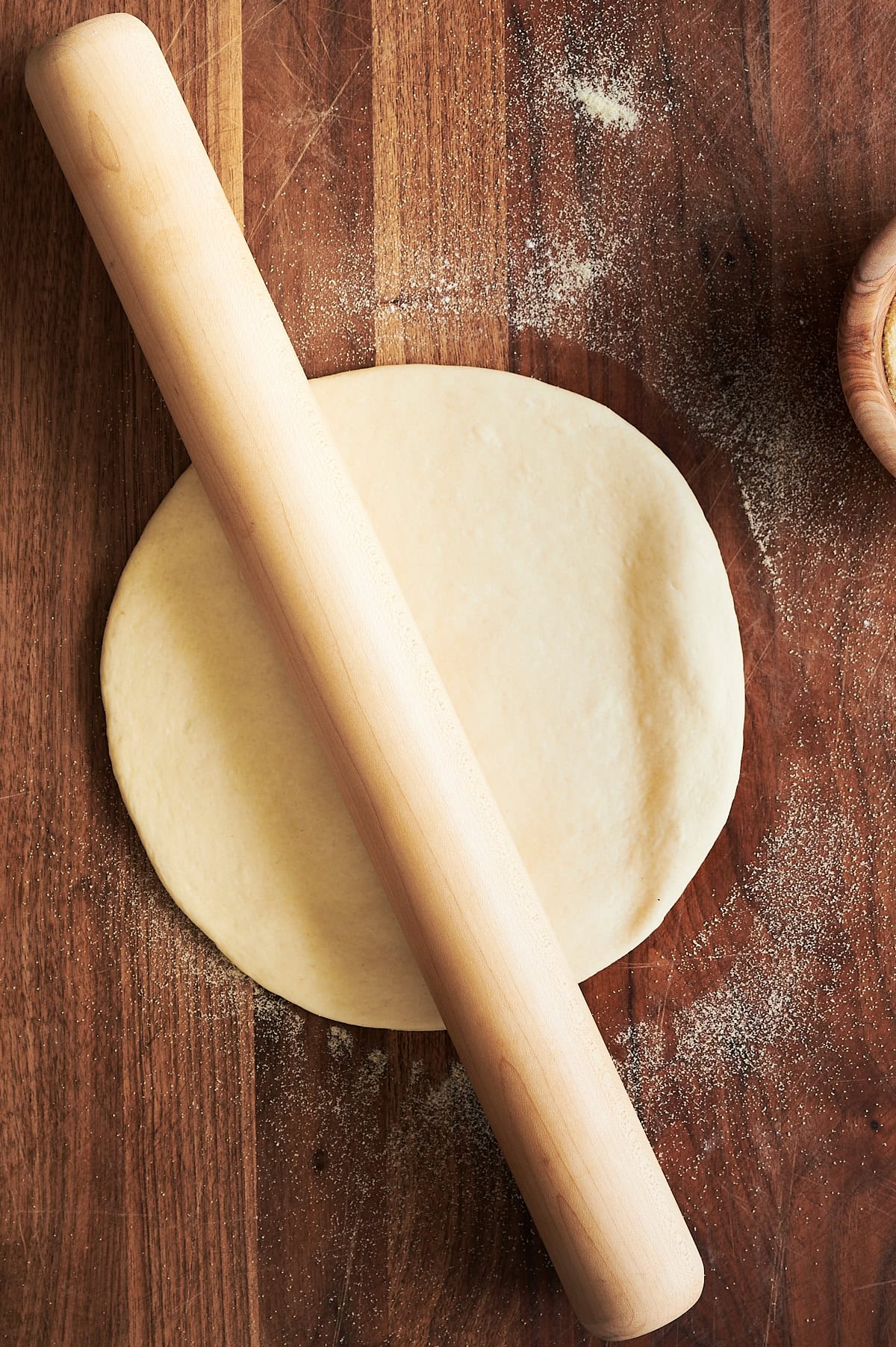 Thin crust pizza dough being rolled out by a French rolling pin angled left on a wooden cutting board.