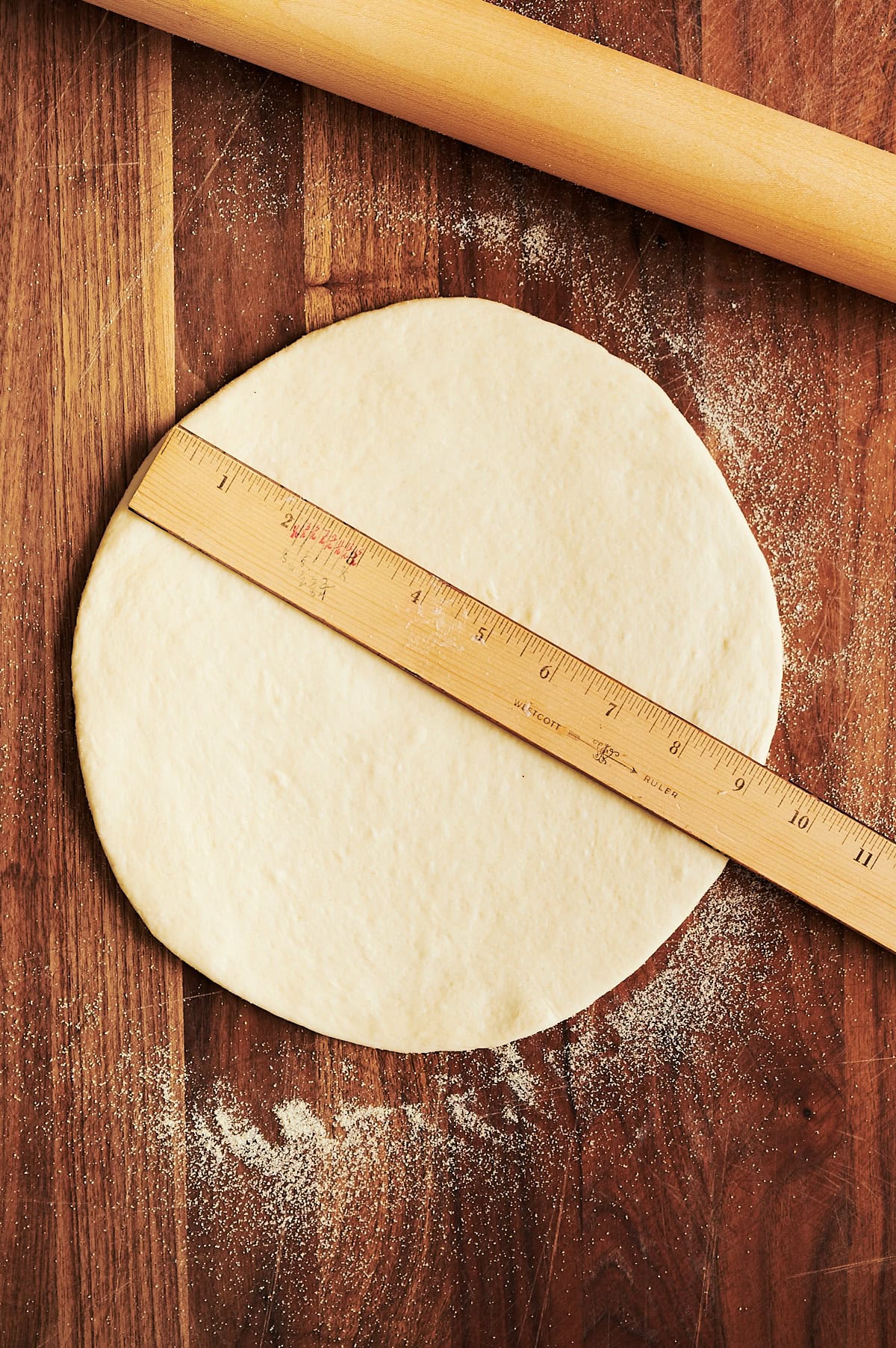 Wooden cutting board with pizza dough rolled out to 9 inches measured by a ruler.