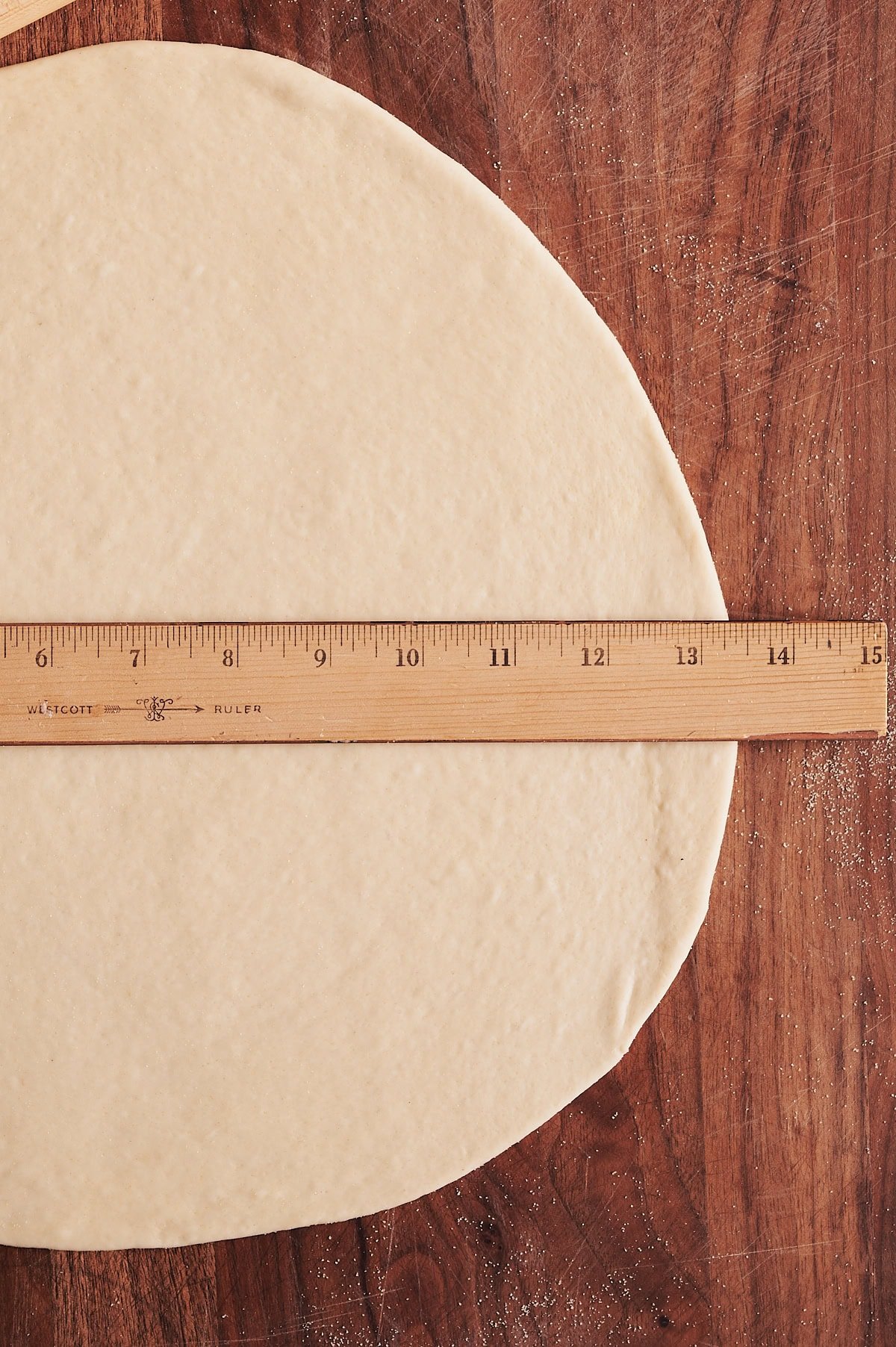 Thin crust pizza dough rolled out on a wooden board rolled to almost 14" in diameter measured by a ruler.