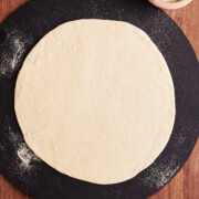 Rolled out thin crust pizza dough on a black pizza stone and wooden cutting board.