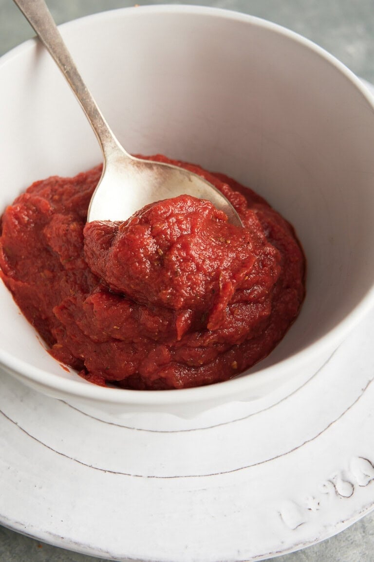 Silver spoon holding tomato paste pizza sauce in a white bowl on a white plate.