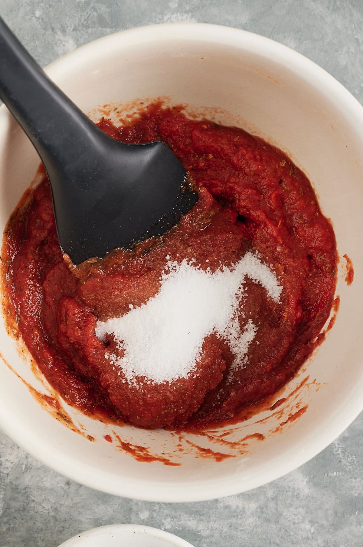 white bowl with tomato paste, seasoning and sugar being stirred with a black spatula.
