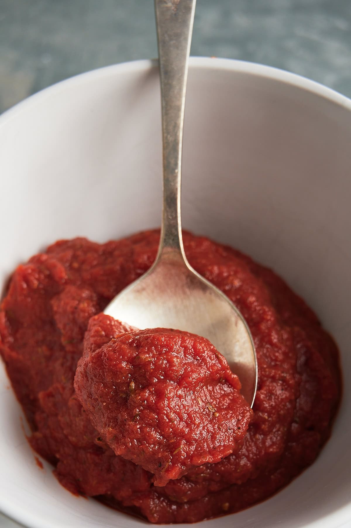 Spoon of tomato paste pizza sauce sitting at an angle in a white bowl of pizza sauce.