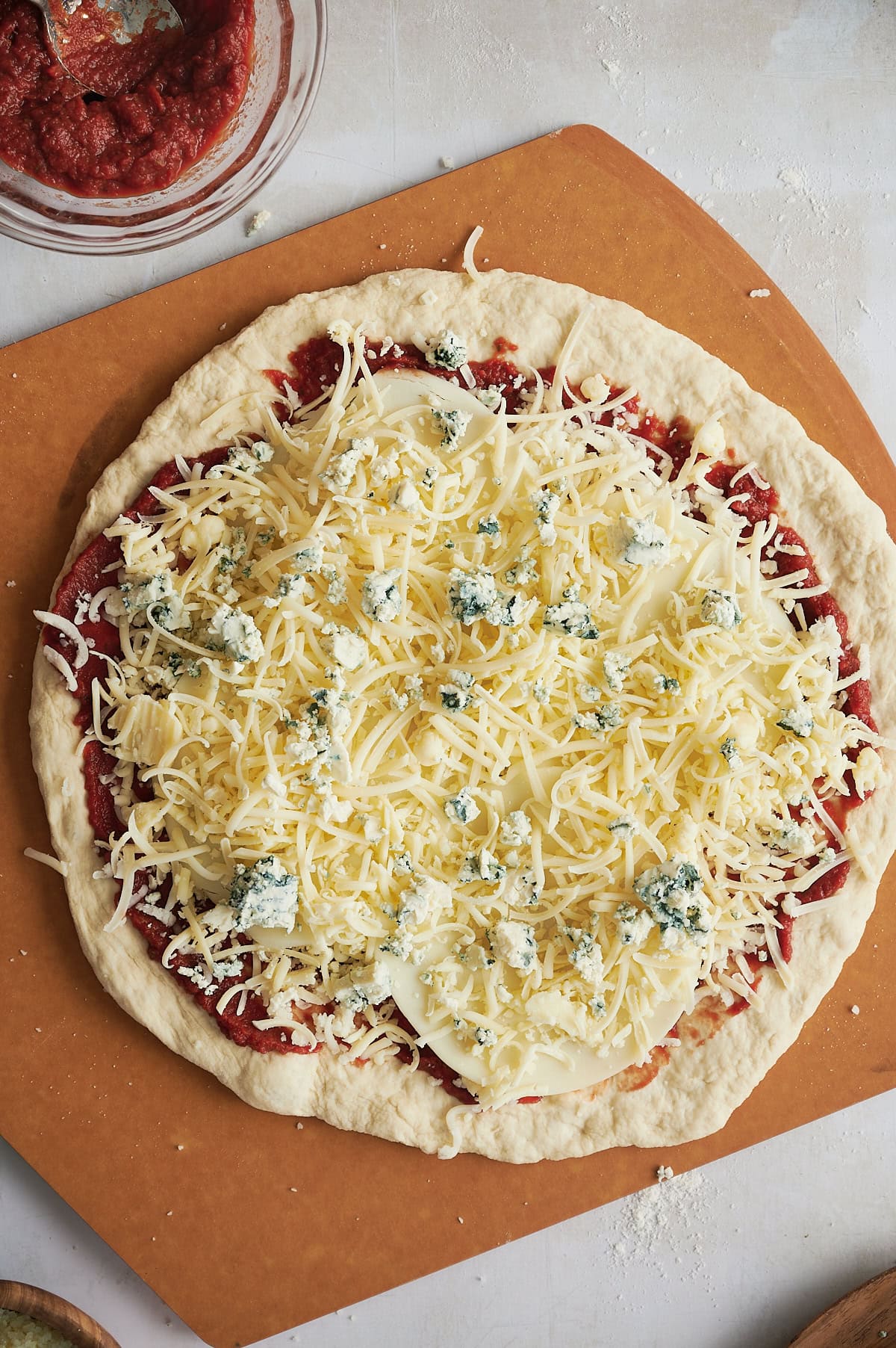 Blue cheese crumbled on an uncooked pizza topped with lots of cheese sitting on a pizza peel.