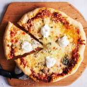 A quarter of a 7 cheese pizza cut out from the whole pie on a wooden cutting board with bowls of ricotta and parmesan cheese.