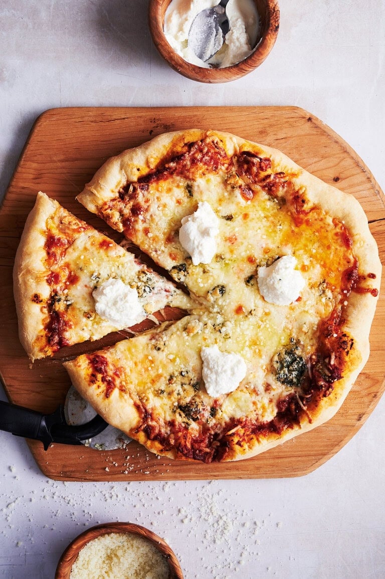 A quarter of a 7 cheese pizza cut out from the whole pie on a wooden cutting board with bowls of ricotta and parmesan cheese.