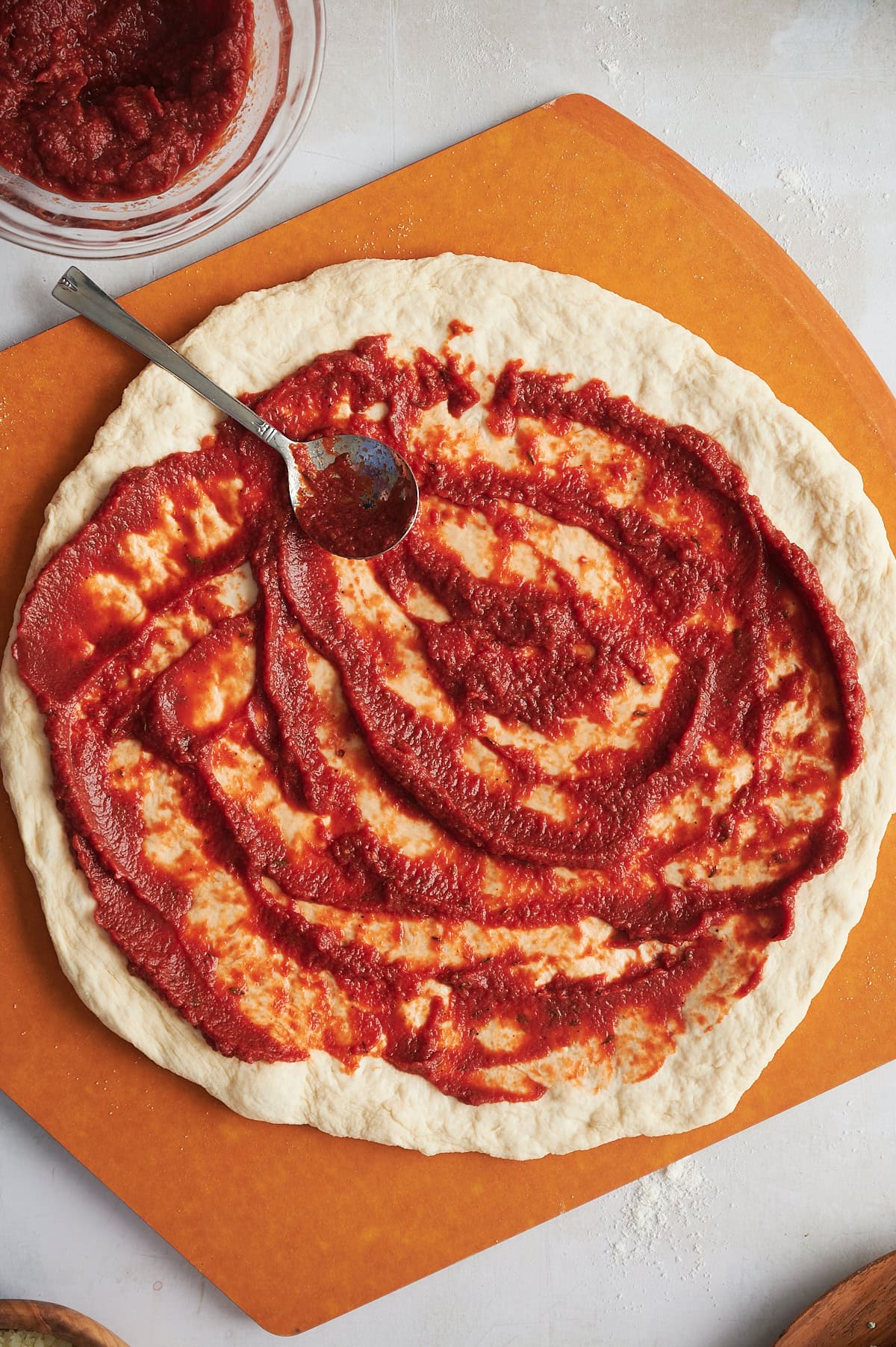 Spoon spreading pizza sauce on pizza dough sitting on a brown pizza peel and bowl of sauce.