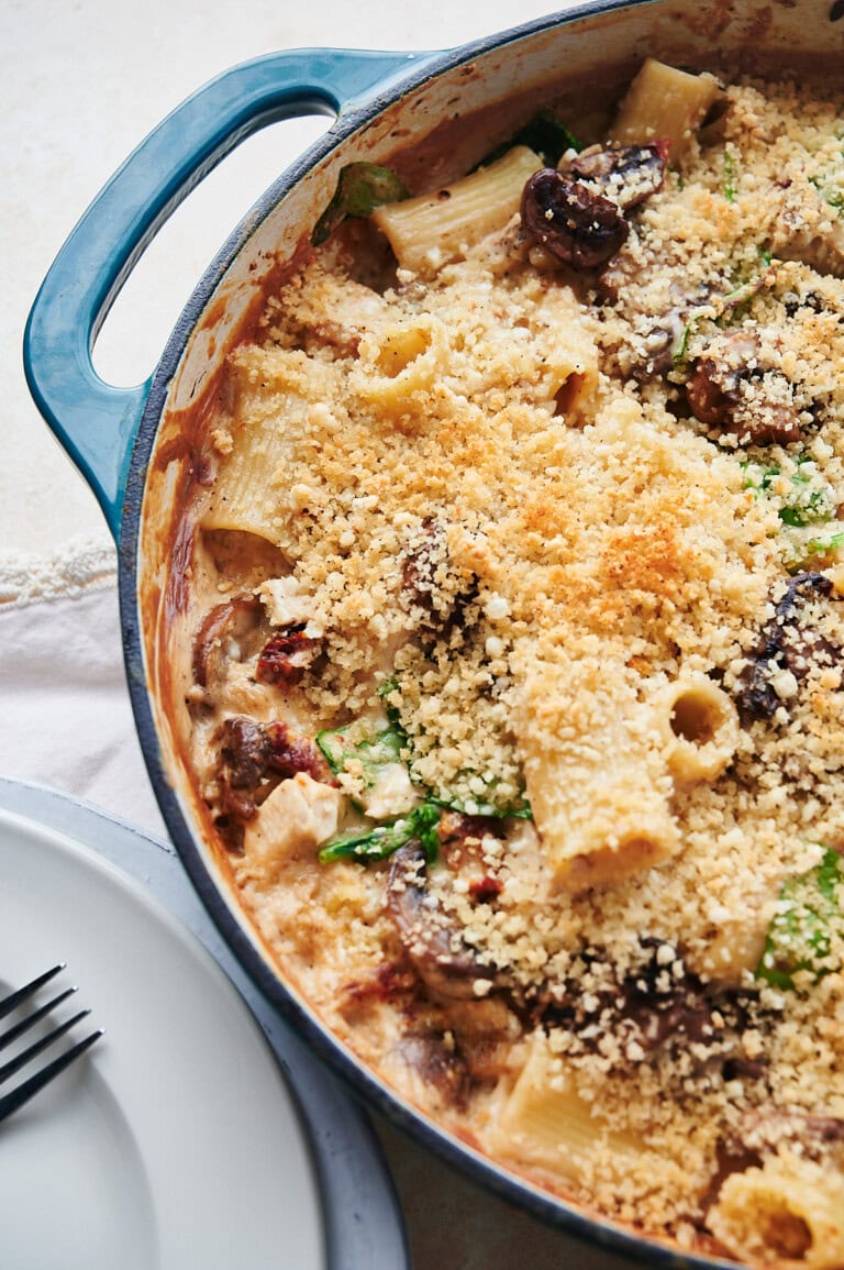 Baked white and teal pan of creamy chicken pasta with mushrooms with a fork and plates on a white lace napkin.