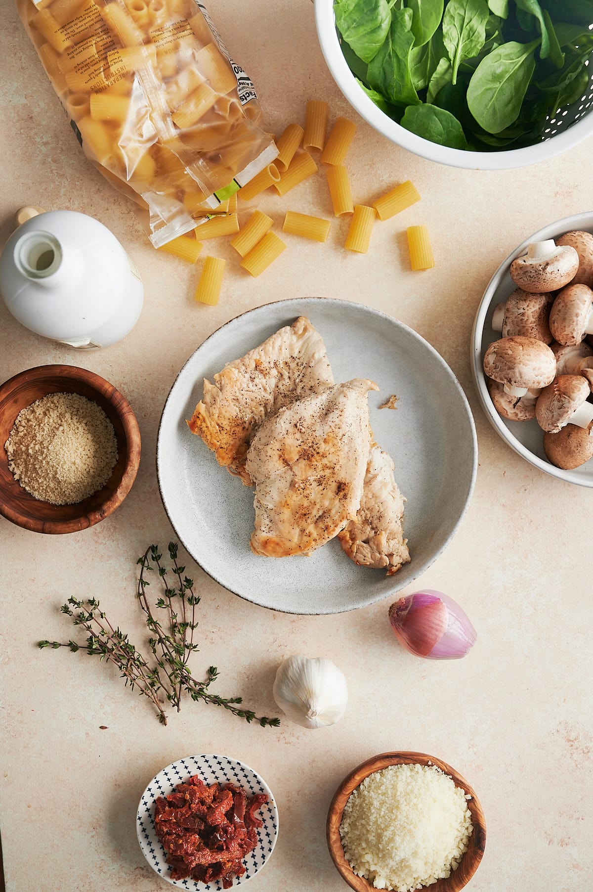 Ingredients for baked chicken pasta on a cream background including chicken, spinach, sun dried tomatoes, parmesan cheese and mushrooms in bowls.