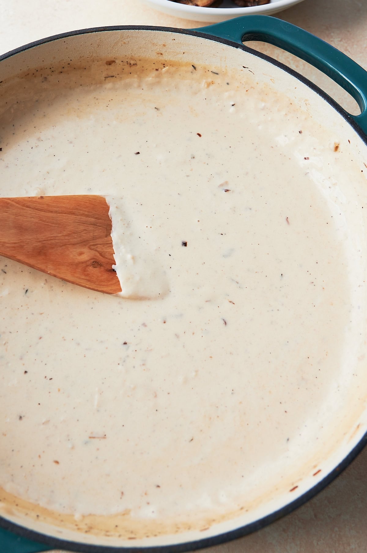Creamy white sauce being stirred by a wooden paddle in a white and teal enamel pan.