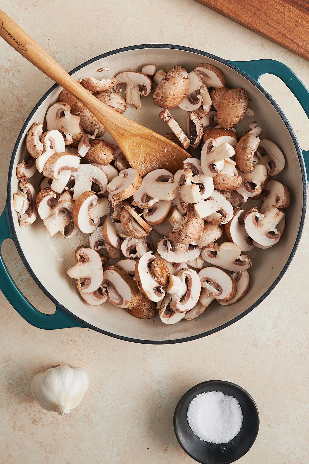 White pan of sauteed mushrooms being tossed with salt by a wooden spoon with head of garlic nearby.