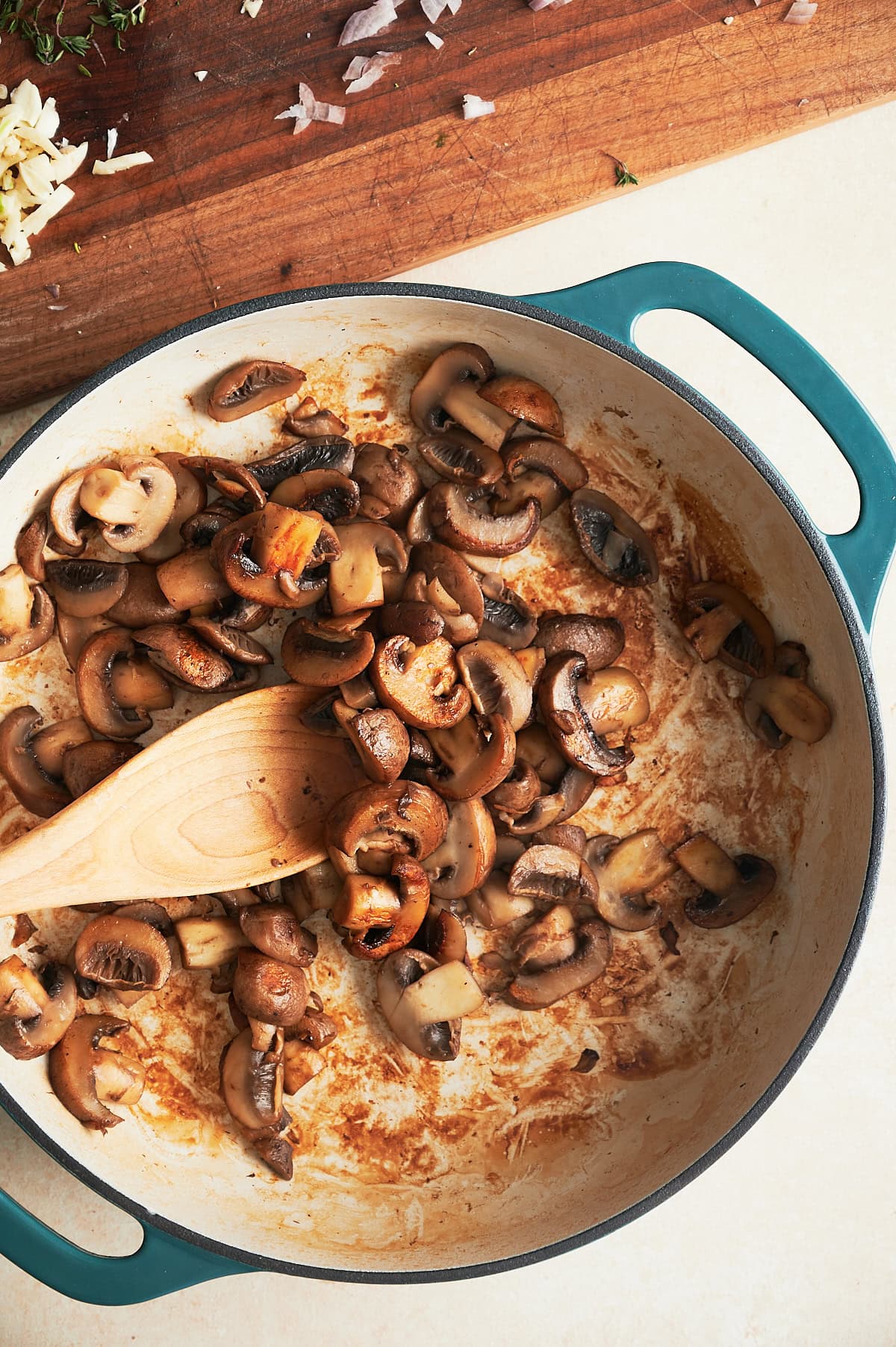 Golden brown mushrooms cooked in a white and teal enamel pan and being stirred by a wooden spoon.