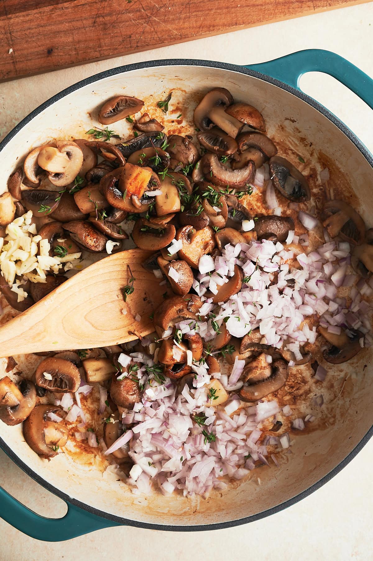 White pan of mushrooms, shallots, garlic and thyme being stirred by a wooden spoon.