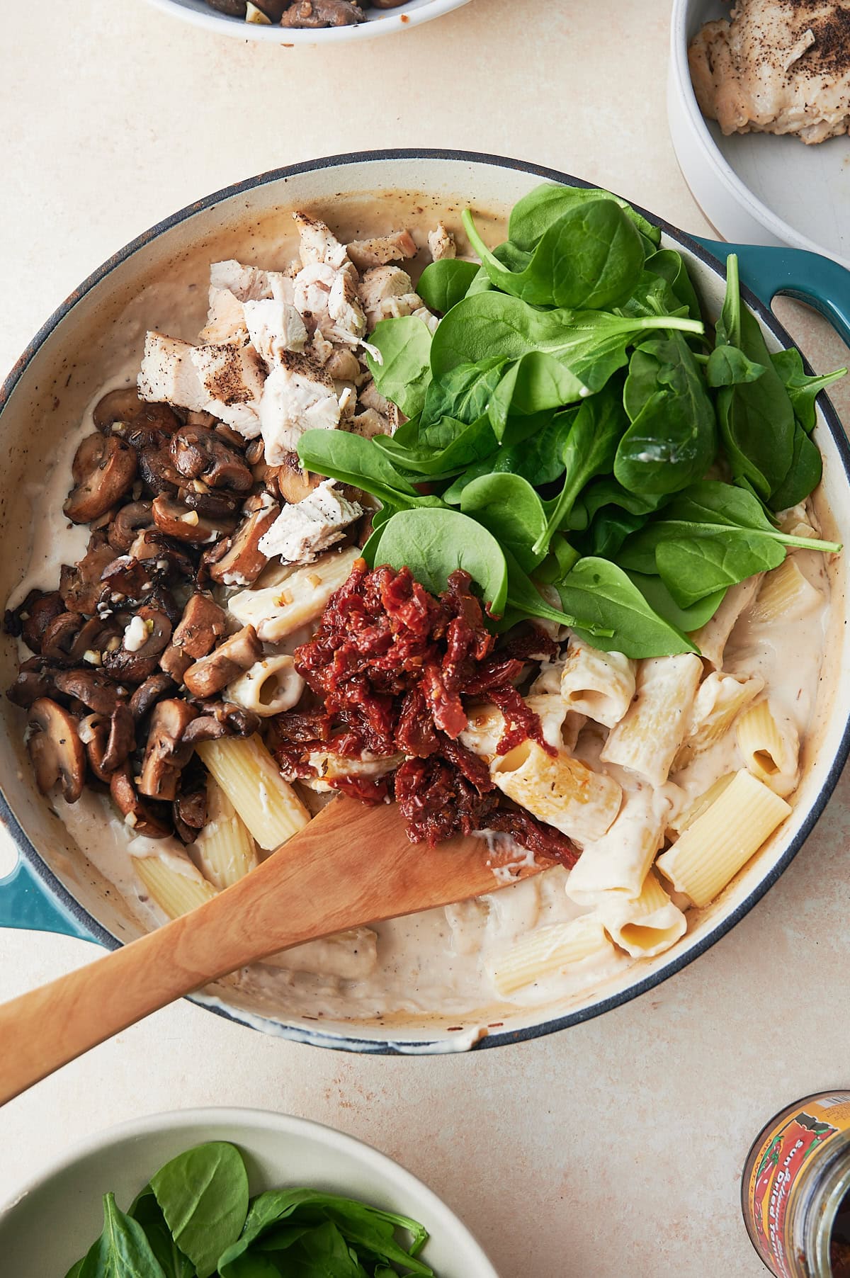 Wooden spoon stirring baked creamy chicken pasta in white sauce in a white and teal skillet.