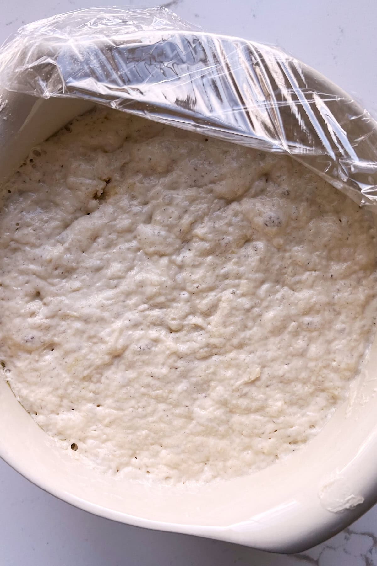 White dough of proofed cinnamon focaccia bread dough partially covered with plastic wrap.