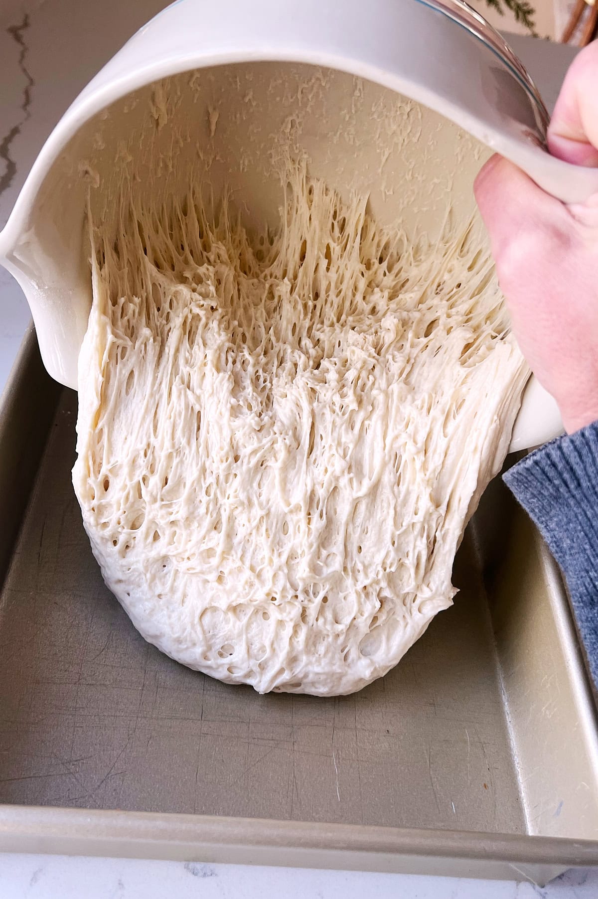 Focaccia dough being poured into gold baking pan for second proofing.