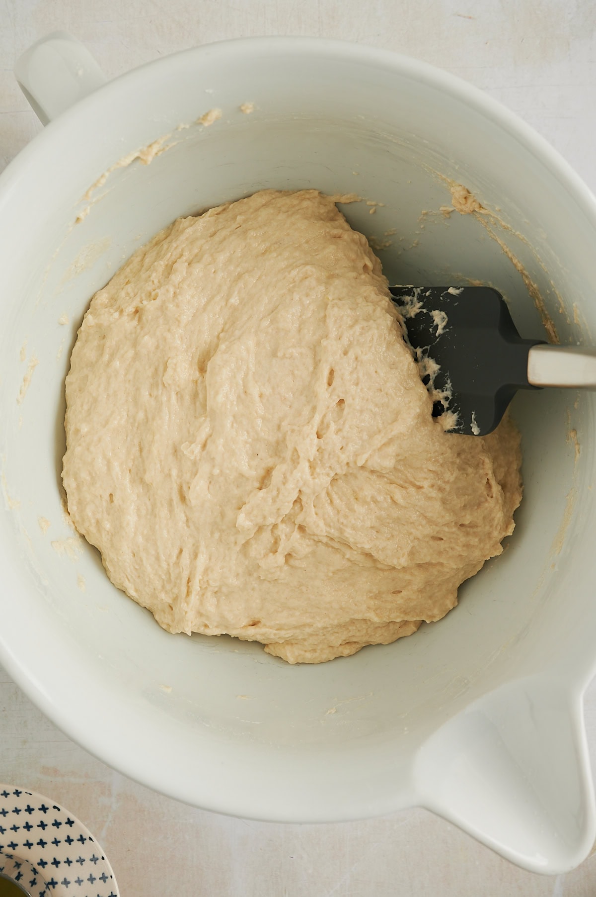 White bowl of cinnamon roll focaccia dough being mixed with black and silver spatula.