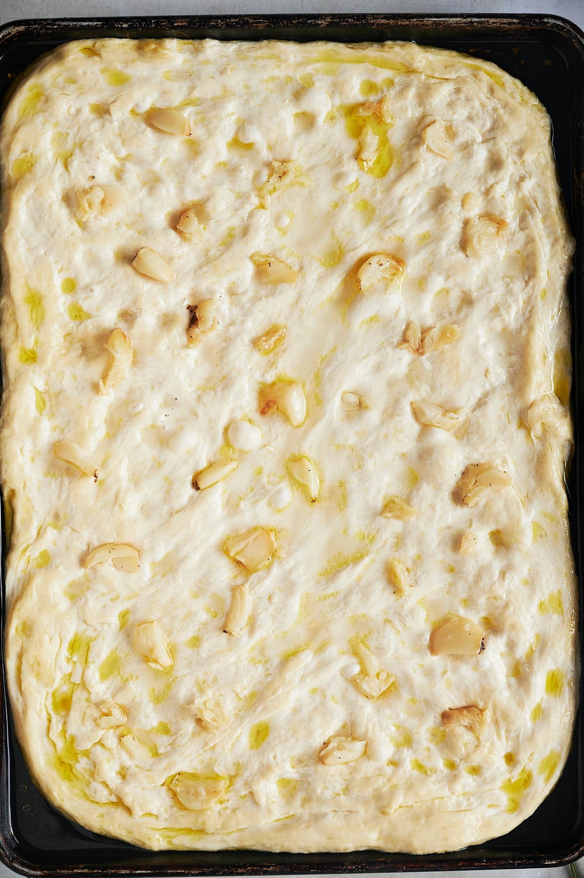 Unbaked second proof garlic topped focaccia bread in black sheet pan.