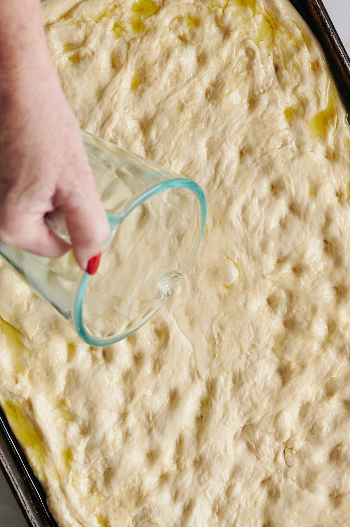 Hand pouring water over dimpled focaccia bread in sheet pan.